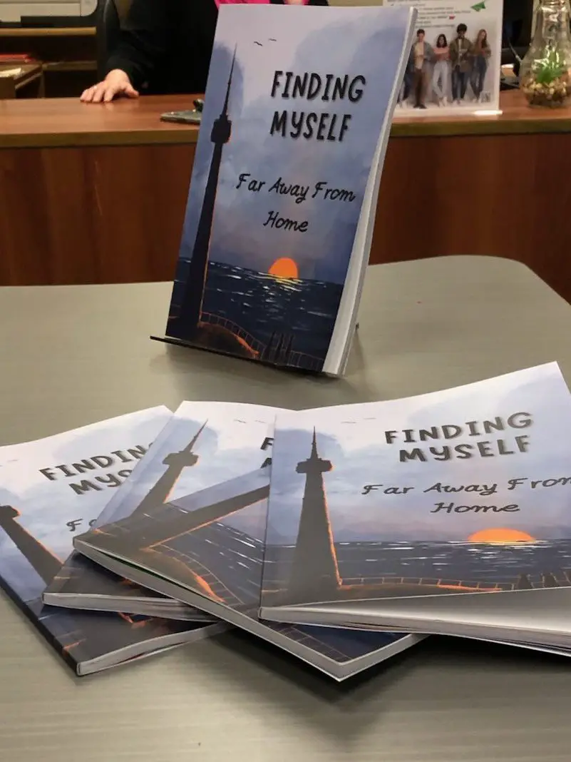 Picture of the book "Finding Myself Far Away From Home" - 1 copy is on a book stand, 4 more are fanned out on a desk.